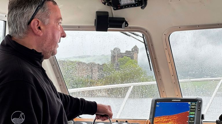 26 August 2023, Great Britain, Drumnadrochit: Skipper Ali Matheson stands on the bridge of a boat on Loch Ness, the ruins of Urquhart Castle can be seen in the background. In Scotland on Saturday began what is believed to be the largest search for the Loch Ness monster, called Nessie, in decades. Dozens of volunteers from around the world posted on Saturday morning in partly pouring rain at 17 observation posts around the famous lake in the Highlands. During the day, boats with special technolog