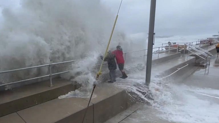 Strong waves hit North Wildwood&#39;s sea walls in New Jersey. People hold on to railings trying to remain upright as they are hit with strong torrents off water.