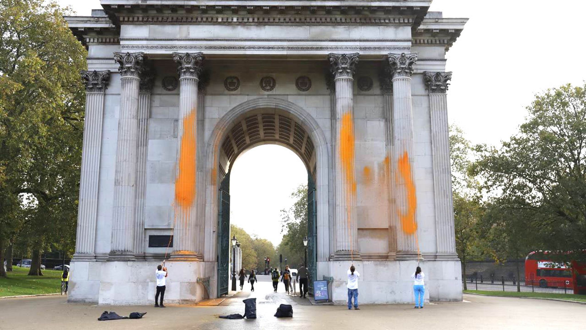 The image shows Wellington Arch in London with bright orange paint sprayed across its white stone columns and facade. Several people in white shirts stand near the monument, with bags on the ground nearby. The classical arch structure with Corinthian columns is surrounded by trees and parkland, with a red double-decker bus visible in the background.