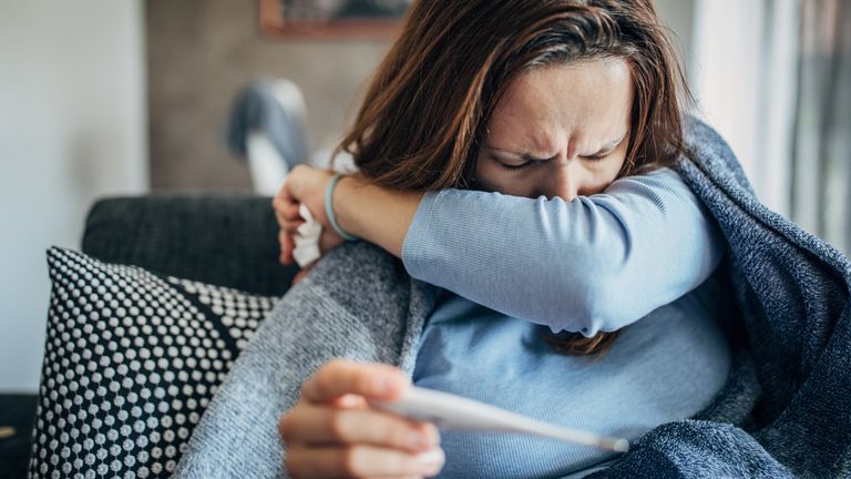 One woman, young woman with Covid-19 symptoms sitting on sofa at home, holding thermometer. Pic: iStock