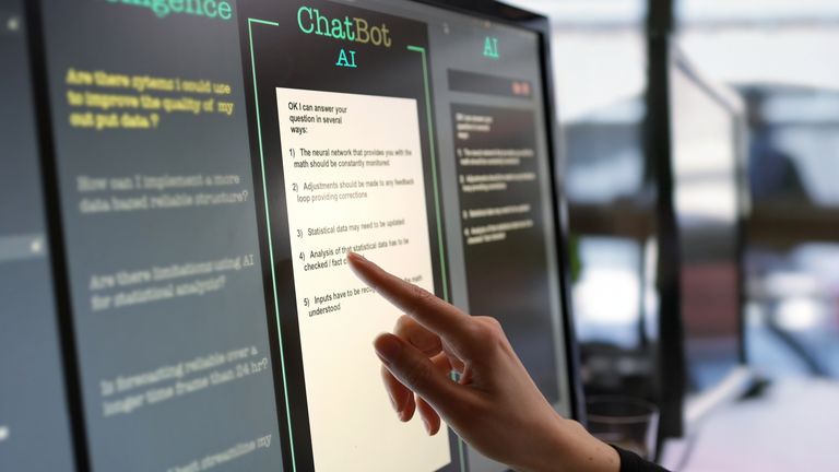 Fotografia em close mostrando um monitor touchscreen sendo usado em um escritório de plano aberto.  A mão de uma mulher fazendo perguntas pré-digitadas para um chatbot de IA e o site de inteligência artificial fornecendo as respostas.