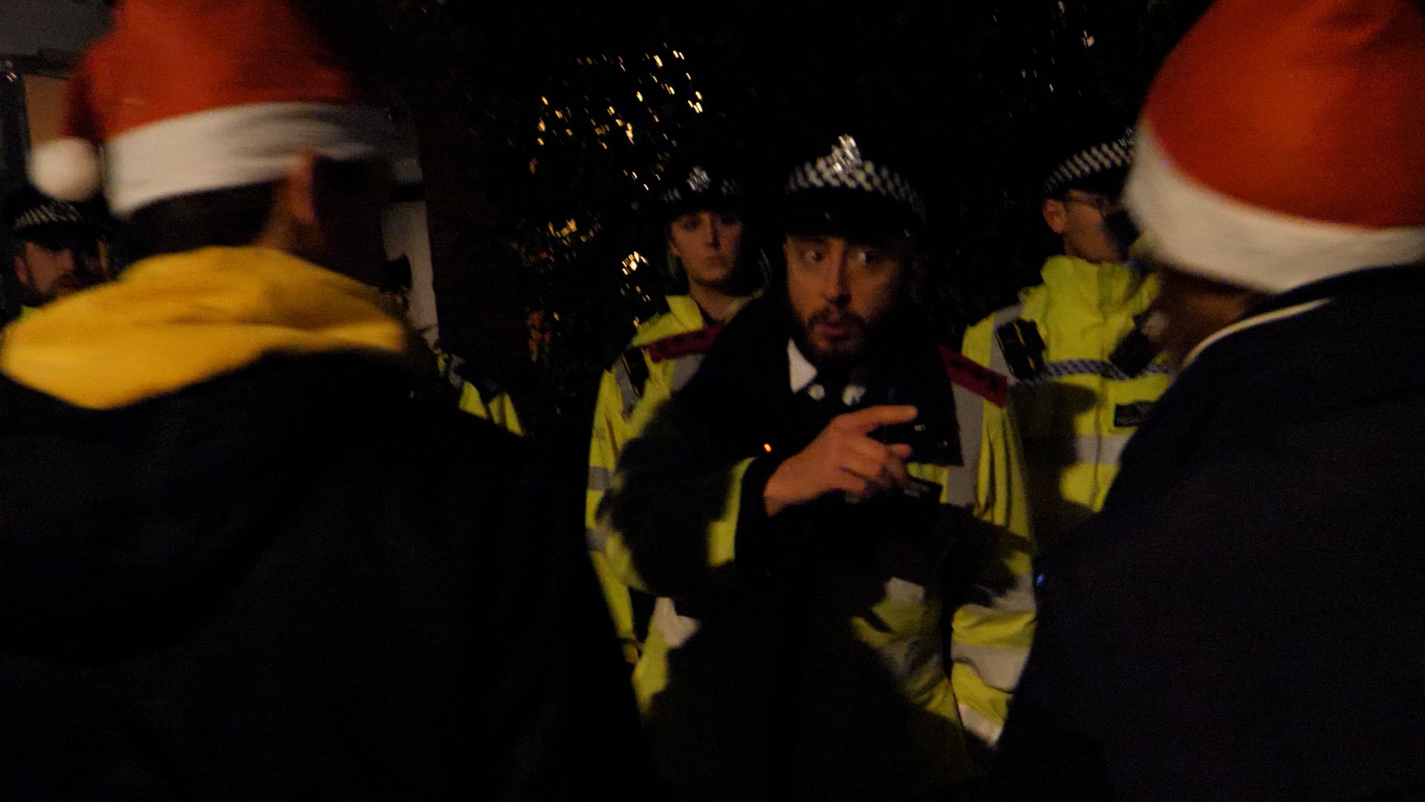 The image shows several police officers in high-visibility yellow jackets and helmets during a nighttime scene. There are blurred Santa hats visible in the frame, suggesting this occurred during the Christmas period. The officers appear to be managing or responding to a situation, with one officer prominently featured in the center of the frame. The scene is dimly lit with some background lighting visible.