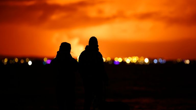 People watch as the night sky is illuminated caused by the eruption of a volcano on the Reykjanes peninsula of south-west Iceland seen from the capital city of Reykjavik, Monday Dec. 18, 2023. (AP Photo/Brynjar Gunnarsson)