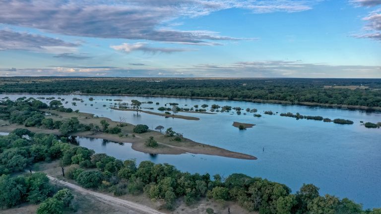 Beautiful view of submerged trees and islands in the Zambezi River in Zambia with gravel road on river bank and clouds in blue sky on far horizon.

