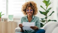 Pic: iStock. For Money blog
Portrait of a happy Afro-American woman sitting on a sofa and looking at a paper in her hand. She is wearing glasses. She might be working from home or studying, or paying a utility bill. She is wearing a green shirt and jeans and there is a laptop in front of her.