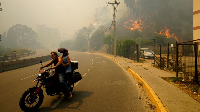 Residents evacuate on a motorcycle as smoke caused by forest fires fill the sky and flames spread into Vina del Mar, Chile, Saturday, Feb. 3, 2024. Officials say intense forest fires burning around a densely populated area of central Chile have left several people dead and destroyed hundreds of homes. (AP Photo/Esteban Felix)
