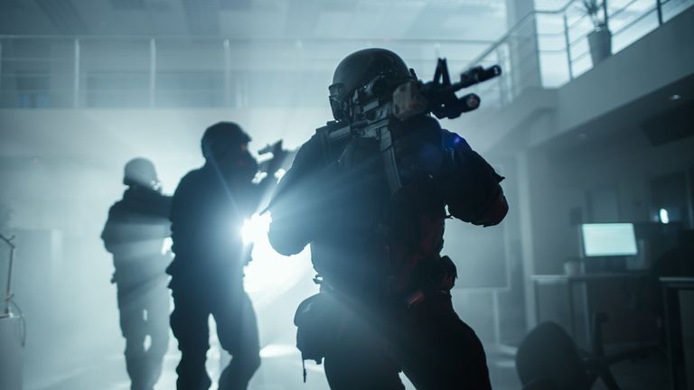 Masked and armed SWAT teams move through the lobby of a dark, occupied office building with desks and computers. Soldiers carrying rifles and flashlights monitor and cover their surroundings.stock