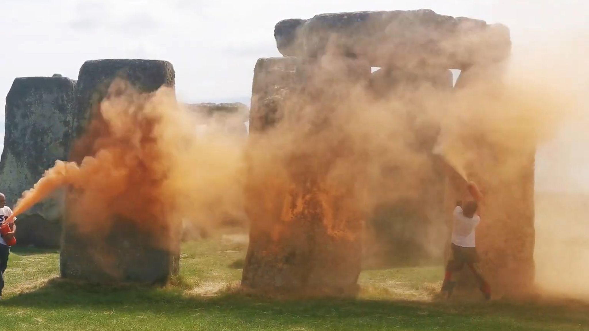 The image shows Stonehenge's ancient stone circle partially obscured by billowing orange powder or paint clouds. Several people are visible near the megalithic stones as the orange substance disperses through the air. The prehistoric monument's distinctive trilithons (two vertical stones topped by a horizontal lintel) are visible through the orange haze on grassy ground under an overcast sky.