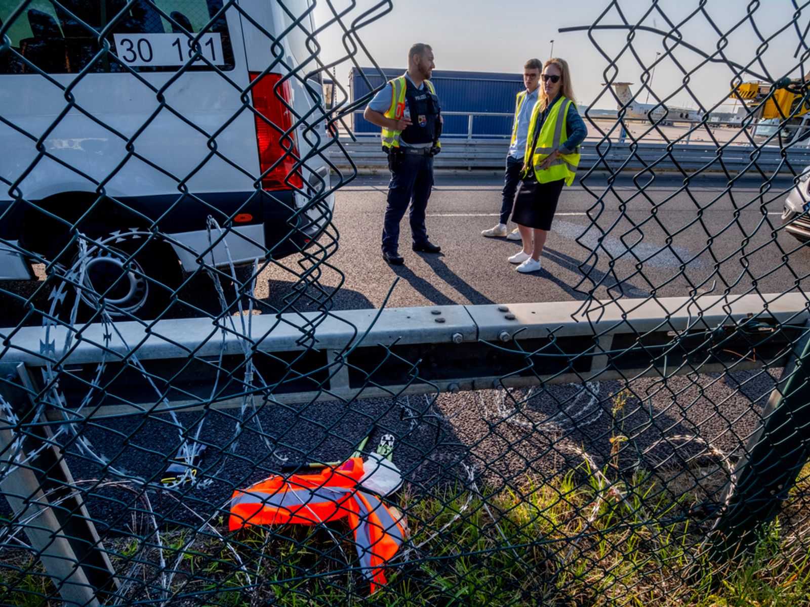 The image shows a damaged chain-link fence with a large hole cut through it. Beyond the fence, three people in high-visibility yellow vests stand on an airport tarmac near aircraft. In the foreground, orange and white safety equipment lies scattered on the ground near the fence breach. The scene appears to be at an airport facility with planes visible in the background.
