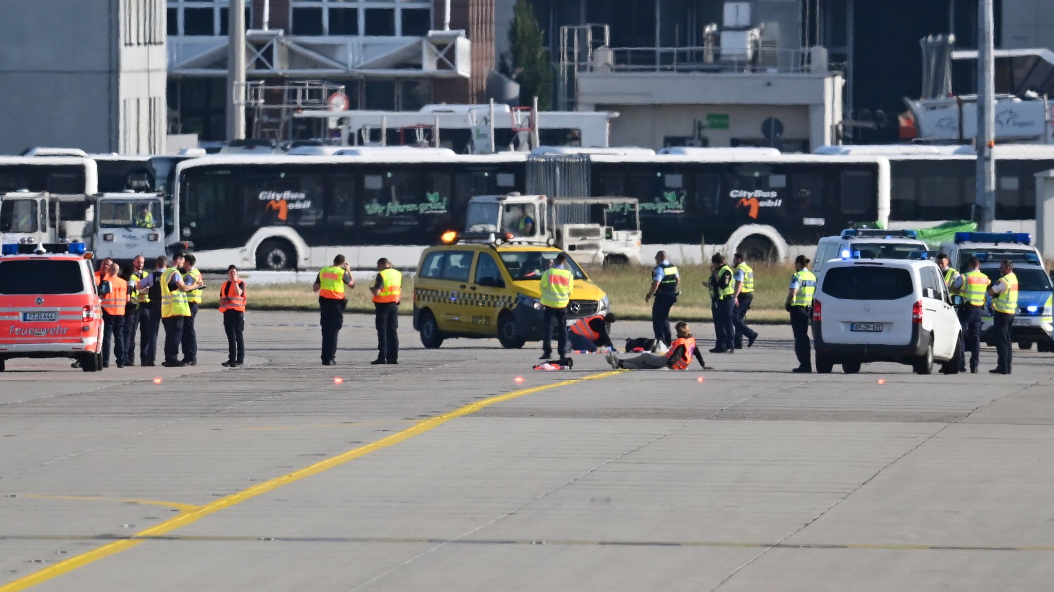 The image shows emergency services personnel in high-visibility vests gathered on an airport tarmac around protesters who appear to be lying on the ground. Multiple emergency vehicles are present, including what appears to be a fire truck and police vans. Airport buses are visible in the background near terminal buildings. The scene takes place on the runway with yellow taxiway markings visible on the concrete surface.