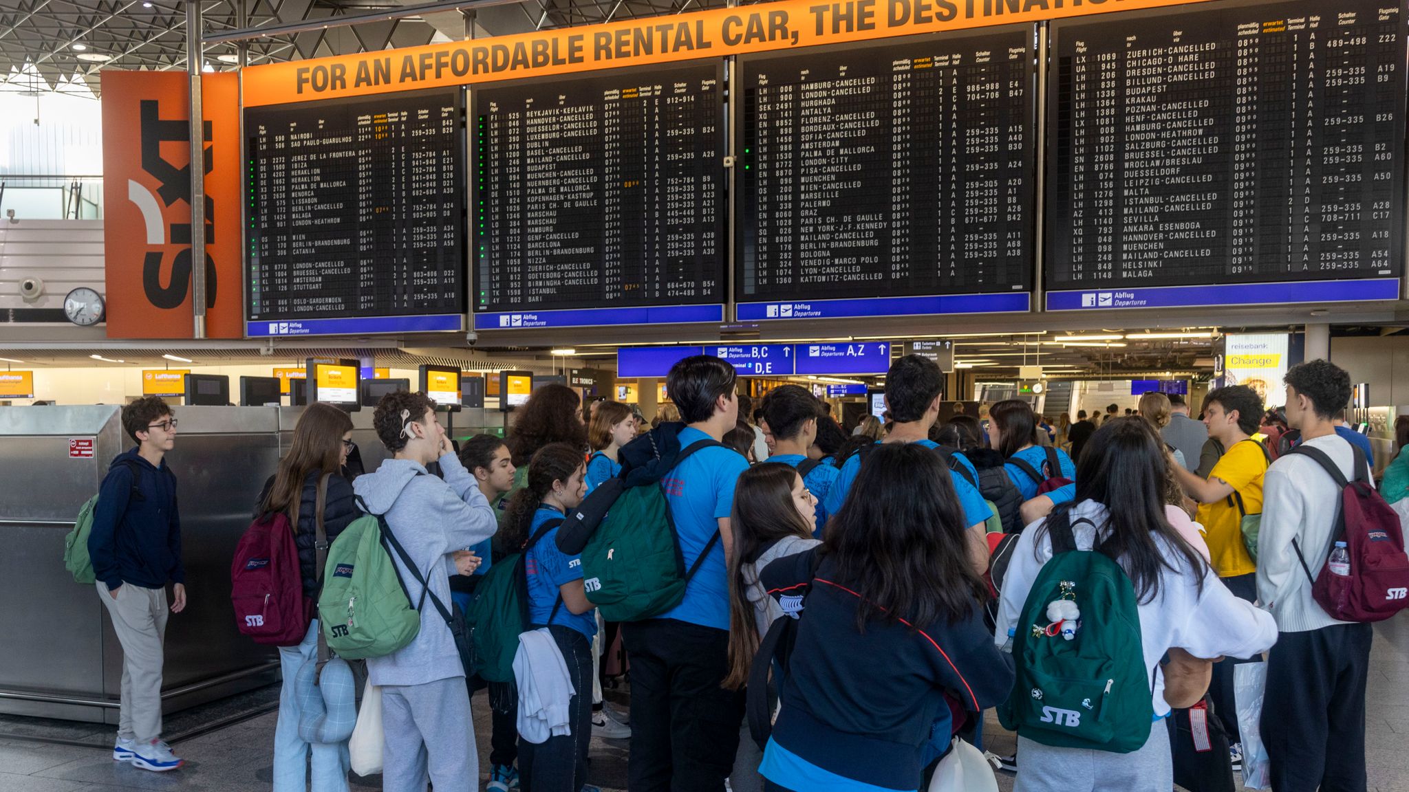The image shows a crowded airport terminal with dozens of passengers gathered around flight information displays. Large electronic boards show numerous flight cancellations and delays, with many entries marked 