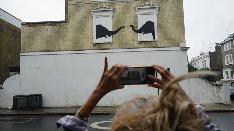 Una mujer se detiene para fotografiar una nueva obra de arte que representa a dos elefantes asomando la cabeza por las ventanas con barrotes abiertas por Banksy en el costado de un edificio en el cruce de Edith Grove y Edith Terrace en Chelsea, al suroeste de Londres. Fecha de la película: martes 6 de agosto de 2024.