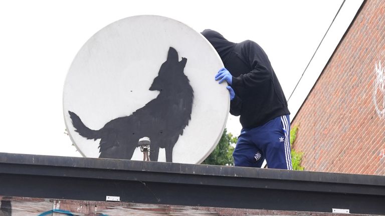 People remove a new artwork by Banksy, depicting a howling wolf painted on a satellite dish that was placed on a shop roof in Peckham, south London. The artist's latest artwork comes a day after he unveiled three monkeys painted on a bridge in Brick Lane, east London. Picture date: Thursday August 8, 2024. PA Photo. See PA story ARTS Banksy. Photo credit should read: Jordan Pettitt/PA Wire