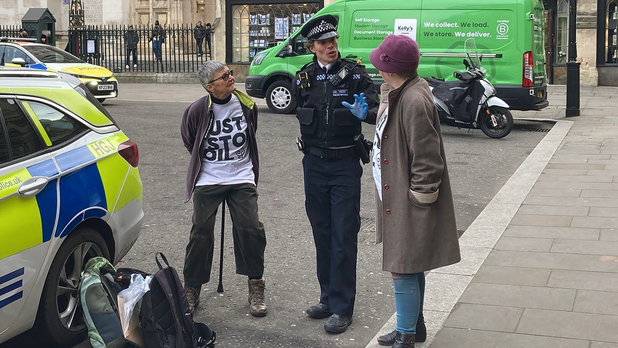 The image shows two Just Stop Oil protesters speaking with a police officer outside Westminster Abbey. One protester wears a 