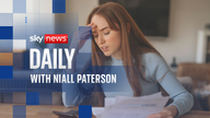 A woman sits at a table, looking stressed as she reviews bills and uses a calculator.