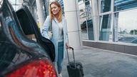 Smiling pleased woman with trolley suitcase closing door of taxicab parked before airport terminal building. Pic: iStock